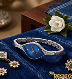 Silver watch with blue face on a blue surface with jewelry and a flower.